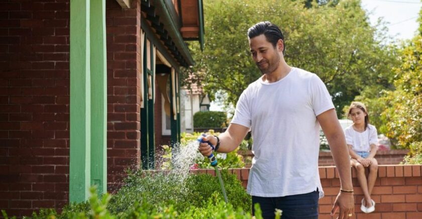 Dad watering garden, while daughter sits on brick fence in background