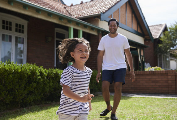 Small girl running and laughing in front garden, dad standing behind her