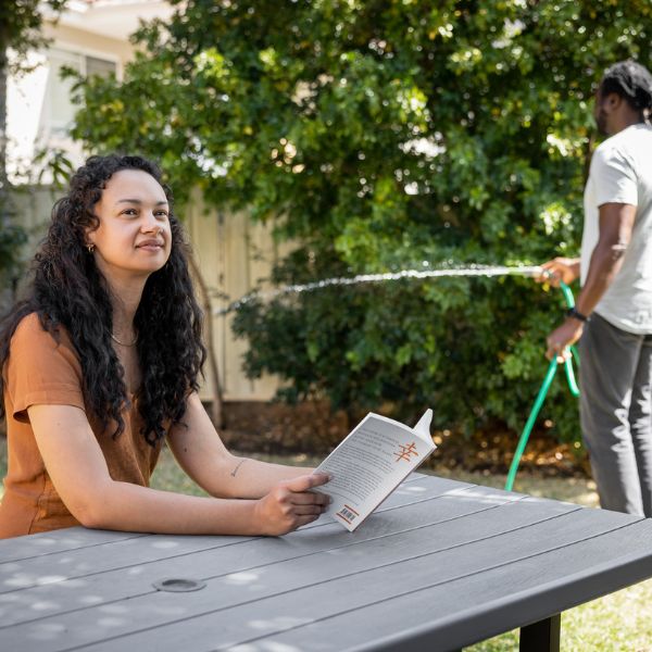 Person reading a book on outdoor bench with another behind watering the garden