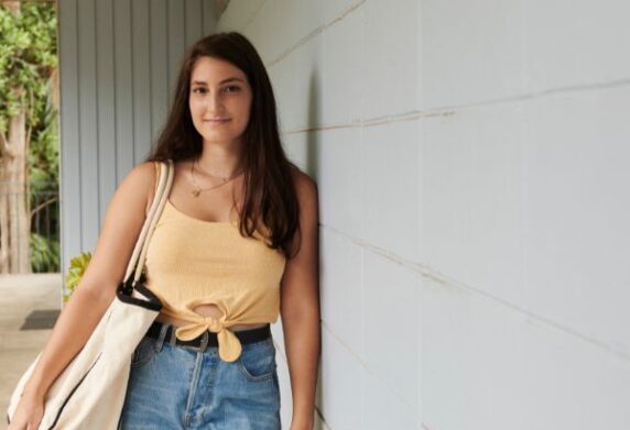 Youth person leaning on wall with bag on shoulder smiling at camera